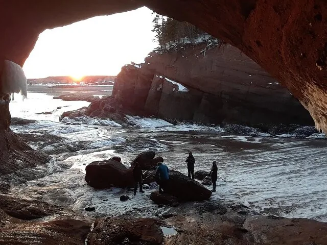 Persone in piedi all'interno di una grande grotta marina che ammirano un tramonto innevato sulla costa.
